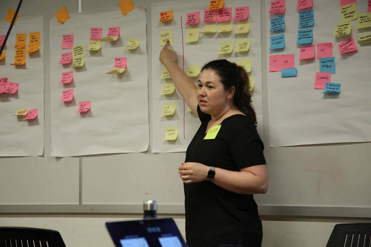a woman giving a talk while pointing to a wall of post-its